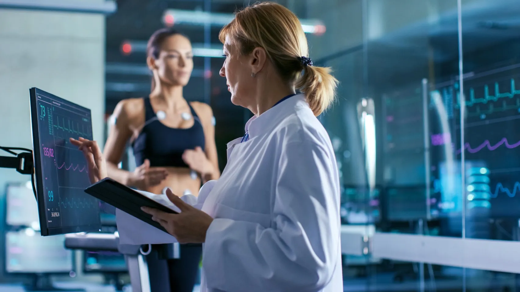 Ärztin mit Tablet am Monitor, dahinter eine Patientin mit EKG-Elektroden beim Belastungstest